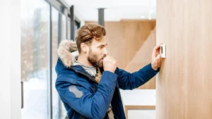 A man in a blue winter coat stands inside, adjusting a thermostat on a wooden wall.