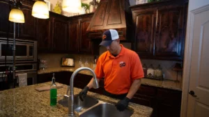 A person wearing an orange shirt and cap stands in a kitchen, closely examining a faucet. Behind them, dark wood cabinets contrast with granite countertops—a space that feels exclusive enough to belong in a high-end membership plan.