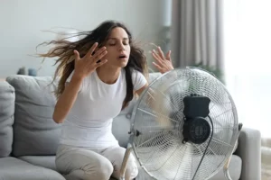 A woman sits on a couch, cooling herself with a fan. Her hair is blowing back, and she has her hands near her face, looking relieved.
