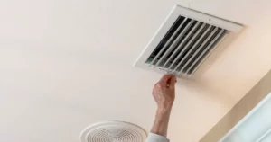 A person adjusts an air vent on a white ceiling, with another circular vent visible nearby.