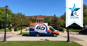 A blue and orange "billyGO" service van is parked on a street in a park area, surrounded by trees. Nearby, a brick building displays a "Reader's Choice Winner" banner, highlighting it as the best air conditioning company around.